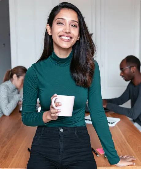 a woman holding a coffee cup in a meeting room with other people sitting at a table in the background.