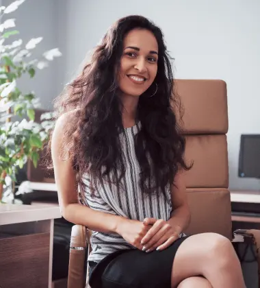 a woman sitting in a chair smiling for the camera with a plant in the background and a computer desk in the background